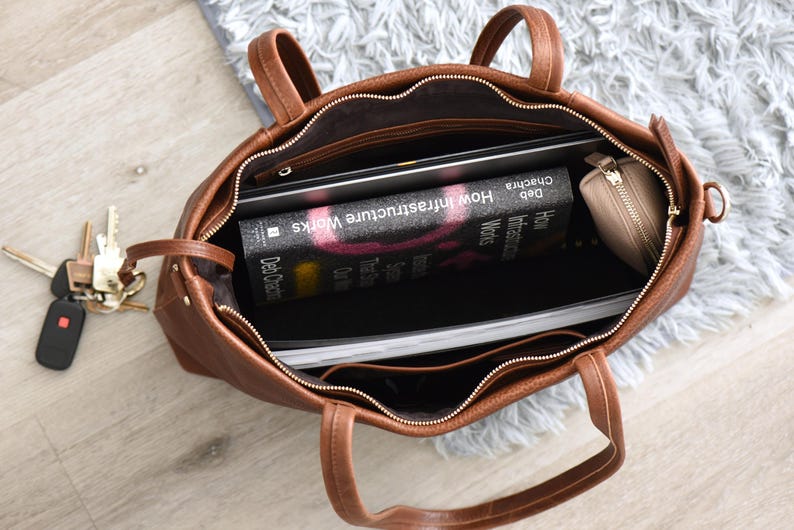 May include: A brown leather tote bag with a zipper closure, containing a laptop, a book titled "How Infrastructure Works," and a small beige wallet. A set of keys with a car remote is visible next to the bag.