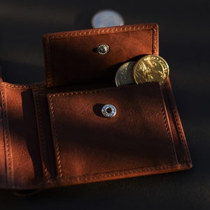 May include: A brown leather wallet with a flap open, revealing a small coin pocket with a few gold coins inside. The wallet has a silver button closure.