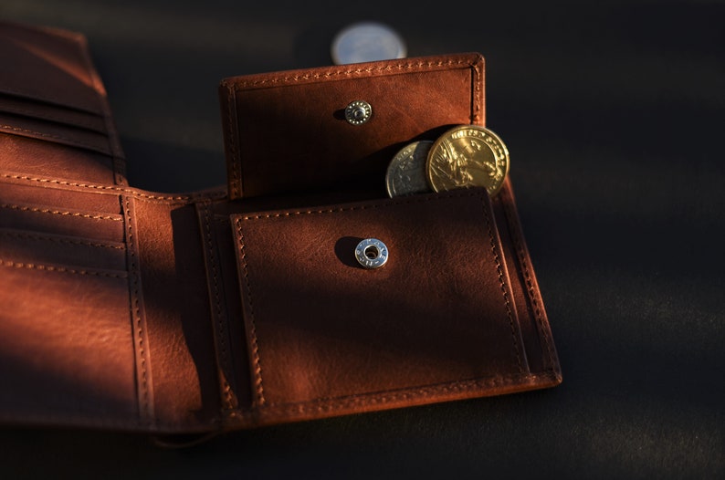 May include: A brown leather wallet with a flap open, revealing a small coin pocket with a few gold coins inside. The wallet has a silver button closure.