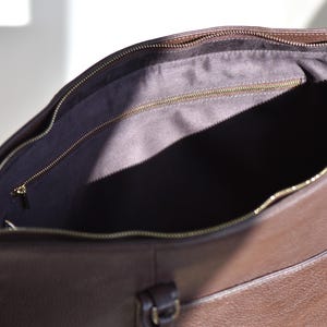 May include: Open view of a brown leather tote bag with a gold zipper. The interior is lined with a dark fabric. The bag has a handle and a small pocket on the front.