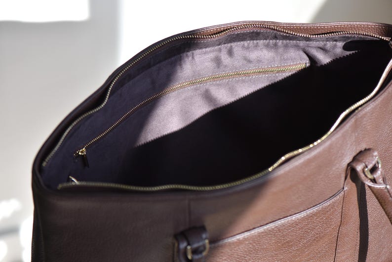 May include: Open view of a brown leather tote bag with a gold zipper. The interior is lined with a dark fabric. The bag has a handle and a small pocket on the front.