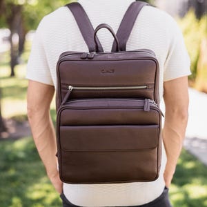 May include: A dark brown leather backpack with a pebbled texture, featuring a top handle, adjustable shoulder straps, and two front zippered compartments. The upper front pocket displays a small embossed logo, and silver-toned metal zippers.
