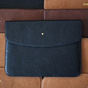 以下が含まれることがあります： 小さな金のハートのディテールが付いた黒い革製のラップトップスリーブ。スリーブは、それぞれ異なるモノグラムが付いた他のいくつかの革製スリーブの上に置かれています。