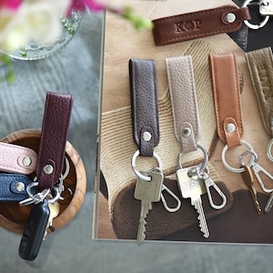 Op de afbeelding: Leren sleutelhangers in verschillende kleuren, waaronder roze, blauw en bruin, met zilveren sleutelringen. Sommige sleutelhangers hebben initialen in reli&euml;f. De sleutelhangers worden getoond in een houten kom en op een patroon oppervlak.