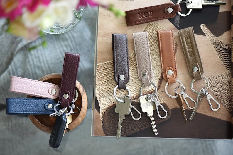 Op de afbeelding: Leren sleutelhangers in verschillende kleuren, waaronder roze, blauw en bruin, met zilveren sleutelringen. Sommige sleutelhangers hebben initialen in reli&euml;f. De sleutelhangers worden getoond in een houten kom en op een patroon oppervlak.