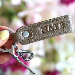 Op de afbeelding: Een taupe leren sleutelhanger met het woord "NAME" in reli&euml;f wordt getoond. De sleutelhanger heeft een zilverkleurige metalen sluiting en sleutelring. De achtergrond is een zachte focus van roze en witte bloemen.