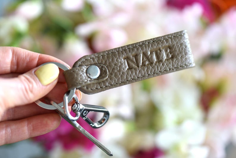 Op de afbeelding: Een taupe leren sleutelhanger met het woord "NAME" in reli&euml;f wordt getoond. De sleutelhanger heeft een zilverkleurige metalen sluiting en sleutelring. De achtergrond is een zachte focus van roze en witte bloemen.