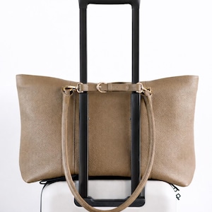 May include: A taupe leather tote bag with a shoulder strap rests on top of a white rolling suitcase. The suitcase has a black handle. The tote bag has gold-colored hardware.