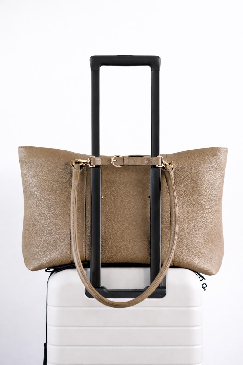 May include: A taupe leather tote bag with a shoulder strap rests on top of a white rolling suitcase. The suitcase has a black handle. The tote bag has gold-colored hardware.
