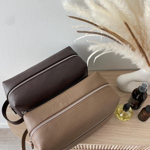 May include: Two leather toiletry bags in shades of brown and tan, with silver zippers and small handles. The bags are rectangular and sit on a light-colored wooden surface. The background includes decorative elements.