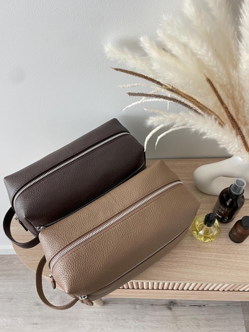 May include: Two leather toiletry bags in shades of brown and tan, with silver zippers and small handles. The bags are rectangular and sit on a light-colored wooden surface. The background includes decorative elements.