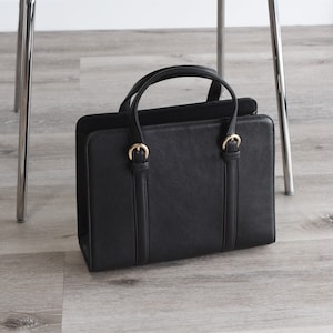 Black leather handbag with gold buckles standing on floor next to chair – professional structured work purse for office.