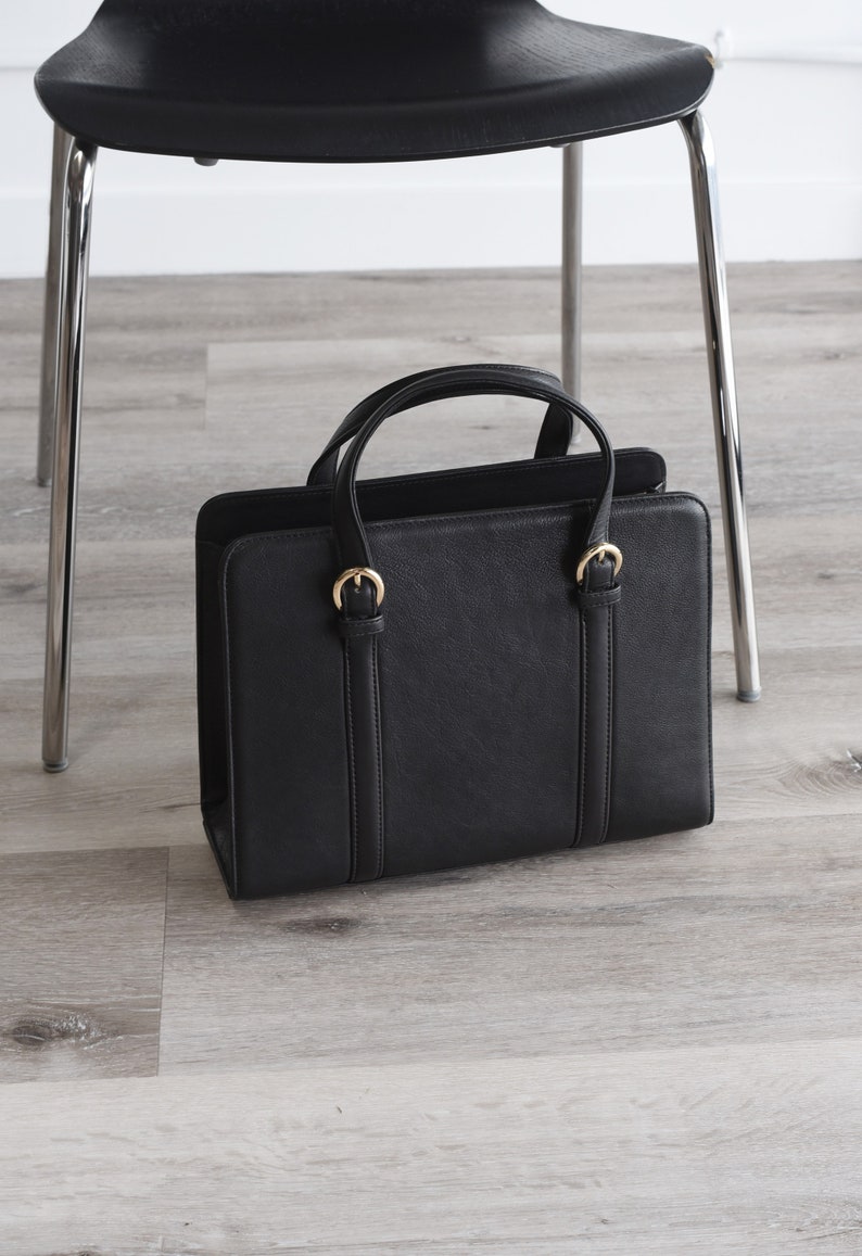 Black leather handbag with gold buckles standing on floor next to chair – professional structured work purse for office.
