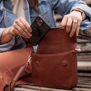 May include: A brown leather crossbody bag with a gold strap and a black phone case with a pop socket inside.