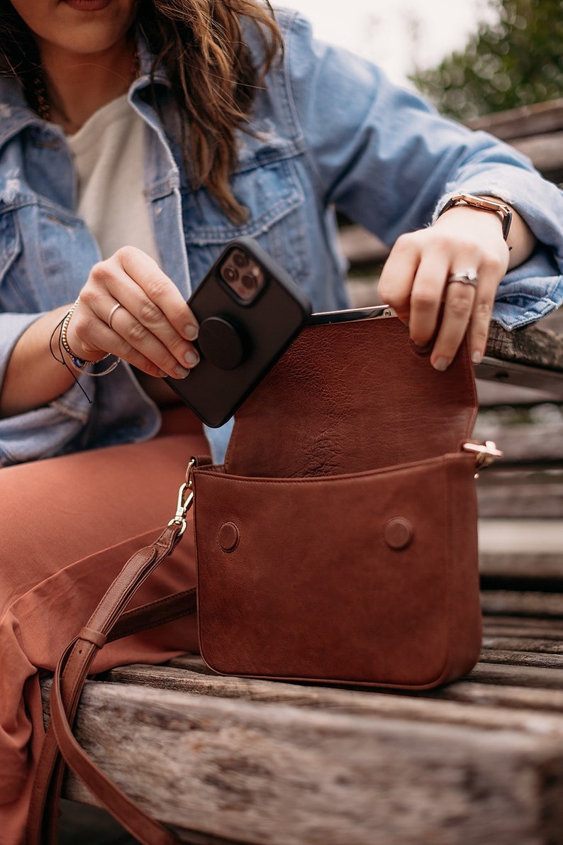 May include: A brown leather crossbody bag with a gold strap and a black phone case with a pop socket inside.