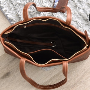 May include: A brown leather tote bag with a top zipper and two handles. The bag has a spacious interior with a dark lining and a small pocket. The zipper is gold-colored. The bag is open, revealing the inside.