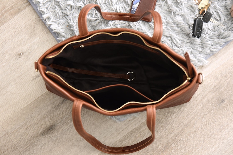 May include: A brown leather tote bag with a top zipper and two handles. The bag has a spacious interior with a dark lining and a small pocket. The zipper is gold-colored. The bag is open, revealing the inside.