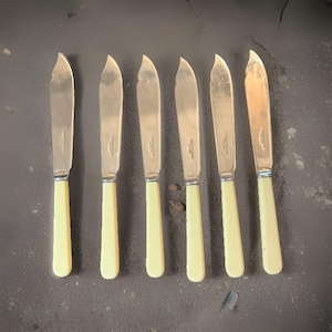 May include: Set of six butter knives with stainless steel blades and cream-colored handles. The knives are arranged in a row, showcasing their classic design. The blades have a slight curve, and the handles are a solid, rectangular shape.
