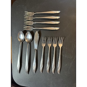 May include: A collection of stainless steel silverware, including forks, spoons, and a butter knife, arranged on a dark surface. The forks have a curved design on the handles. The spoons and knife have a similar design. The silverware set is ideal for dining.