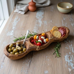 Könnte beinhalten: Ein hölzernes Serviertablett mit drei verbundenen Abschnitten. Ein Abschnitt enthält grüne Oliven, ein anderer Spieße mit Tomaten und Mozzarella, und der dritte Cracker und Salami. Eine silberne Gabel und Rosmarinzweige sind ebenfalls vorhanden.