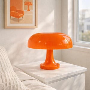 May include: A bright orange mushroom-shaped table lamp with a glossy finish. The lamp sits on a white nightstand. An orange-themed artwork hangs on the wall in the background. The lamp's design is retro and modern.