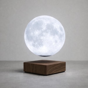 Puede incluir: Lámpara lunar flotante con superficie detallada, suspendida sobre una base cuadrada de madera. La lámpara emite una suave luz blanca, creando un ambiente celestial. La base es de madera oscura.