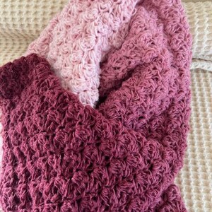 May include: A handmade, textured crochet blanket in shades of pink and burgundy. The blanket features a dense, raised stitch pattern, creating a soft and cozy appearance. The colors transition from light pink to a deep burgundy.