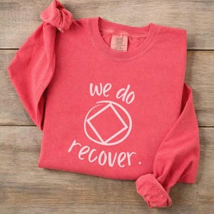 Peut inclure: T-shirt à manches longues corail avec les mots "we do recover" et un symbole blanc de cercle et de losange. Le t-shirt est fait d'une matière douce et a des manches retroussées.
