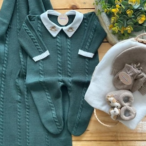 Peut inclure: Ensemble de vêtements pour bébé vert foncé, comprenant une barboteuse en tricot avec un col blanc et une couverture assortie. La barboteuse présente un motif en tricot torsadé et un petit détail de cheval à bascule brodé. Un chapeau, des moufles et des chaussons sont également inclus.