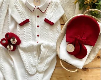 May include: A white cable-knit baby outfit set with red trim, including a romper, hat, and mittens. The set also includes red baby booties with bear faces. The items are arranged on a wooden surface.