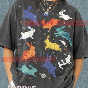 May include: Dark gray t-shirt featuring colorful rabbits, stars, and planets. The design includes rabbits in blue, orange, white, yellow, and teal. The text "BORATICOETSY.COM" is printed across the shirt, and the word "Pepper" is at the bottom.