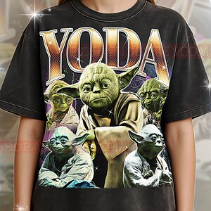 Peut inclure: T-shirt noir avec le mot "YODA" en or et blanc. Le t-shirt présente plusieurs images de Yoda, le Maître Jedi, dans différentes poses. Le design a un aspect vintage et vieilli.