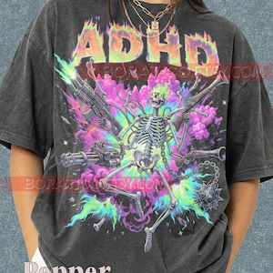 May include: A charcoal gray t-shirt featuring a graphic of a skeleton with multiple arms holding various firearms, including a minigun and rifles, surrounded by vibrant purple and green smoke and flames, with the text ADHD in a colorful font.
