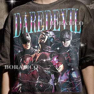 May include: A charcoal gray short-sleeve t-shirt featuring a graphic print of the character Daredevil in multiple poses. The word DAREDEVIL is printed in large, metallic-toned letters at the top, accompanied by the text BORATICO and lightning effects.