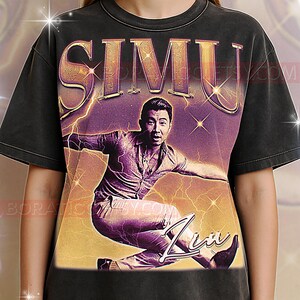 以下が含まれることがあります： ダイナミックなポーズのシム・リウのグラフィックが特徴のダークグレーのTシャツ。名前「SIMU」は大きな金色の文字で表示されています。デザインには稲妻とサインが含まれています。シャツはヴィンテージ感のある、使い古したような外観です。