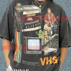 Può includere: T-shirt grigio scuro con una pila di videocassette VHS con titoli di film come "Ritorno al futuro" e "Ghostbusters". Il design include una TV retrò, un videoregistratore e personaggi cinematografici. La parola "VHS" è in grandi lettere arancioni.