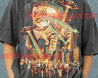Pode incluir: Camiseta cinza escuro com um gato tigrado atirando raios laser em uma cidade. Dois OVNIs disparam raios coloridos. Uma multidão de pessoas foge do gato. A palavra "Pepper" está na parte inferior.