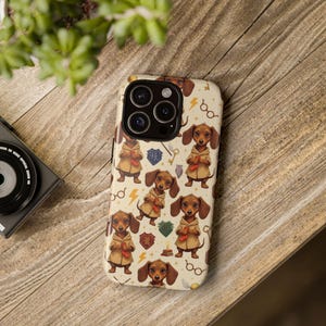 Puede incluir: Una funda de teléfono con un patrón repetido de perros salchicha de dibujos animados con túnicas color canela, sosteniendo pergaminos. El fondo es de color crema con rayos, gafas y escudos. La funda tiene un recorte para cámara de lentes múltiples.