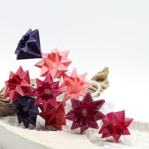 Könnte beinhalten: Eine Lichterkette mit origami-förmigen Sternen in Rosa-, Rot- und Lilatönen. Die Lichter sind in einer weißen, flachen Schale mit einem Stück Treibholz angeordnet. Die Lichter sind an einem weißen Draht.