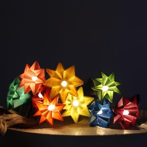 Könnte beinhalten: Eine Ansammlung farbenfroher sternförmiger Lichter, jedes mit einem hellen Zentrum, auf einer Oberfläche angeordnet. Die Lichter sind in den Farbtönen Rot, Orange, Gelb, Grün, Blau und Rosa gehalten. Ein Stück Treibholz befindet sich im Vordergrund.