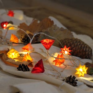 Könnte beinhalten: Eine LED-Lichterkette mit geometrischen, sternförmigen Abdeckungen in Rot-, Orange- und Gelbtönen. Die Lichter leuchten und werfen einen warmen Schein auf verstreute getrocknete Herbstblätter, Tannenzapfen und Eicheln auf einem weißen Stoff.
