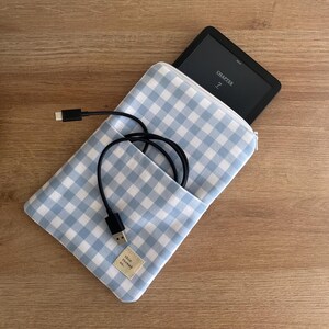 May include: A light blue and white gingham patterned e-reader case with a black charging cable and an e-reader. The e-reader is open to Chapter 7. The case has a small tag that says "this thread co."