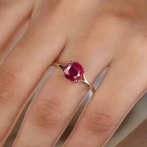 May include: A gold ring featuring a large, oval-cut ruby gemstone. The ruby is a deep red color and is set in a delicate, open-work setting. The ring's band is a thin, polished gold.