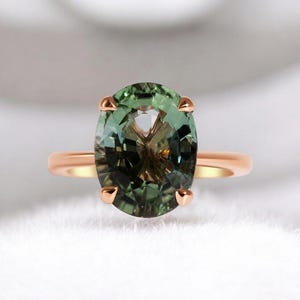 Op de afbeelding: Een roségouden ring met een grote, ovaal geslepen groene edelsteen. De edelsteen heeft een meerkleurig uiterlijk, met tinten groen en bruin. De ring heeft een eenvoudige, gepolijste roségouden band.