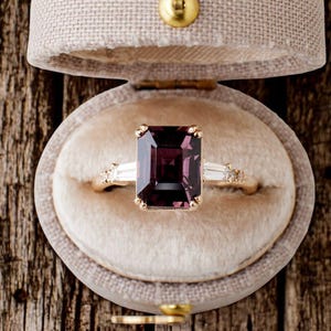 Peut inclure: Une bague en or avec une grande pierre précieuse rectangulaire, de couleur violet foncé. La bague comporte de plus petites pierres de taille baguette claires de chaque côté de la pierre principale. La bague est présentée dans un écrin en velours beige.