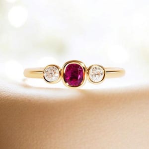 Könnte beinhalten: Ein goldener Ring mit einem zentralen, kissenförmig geschliffenen Rubin und zwei kleineren, runden, klaren Edelsteinen. Der Ring hat ein poliertes Goldband. Die Edelsteine sind in einer Zargenfassung gefasst.