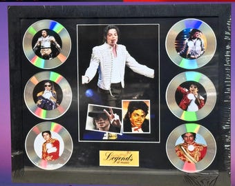 May include: Framed Michael Jackson memorabilia featuring a central photo and smaller images on CD-shaped discs. The central image shows Michael Jackson in a white jacket. The discs display various performance shots. The frame has the text "Legends of music".