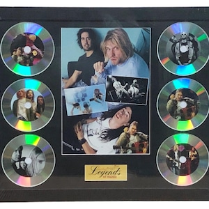 Puede incluir: Enmarcado de recuerdos musicales con un collage de fotos central y seis CD. El collage incluye fotos de la banda e imágenes más pequeñas. Los CD están dispuestos alrededor de la imagen central. Una placa dorada en la parte inferior dice "Legends of music". El marco es negro.