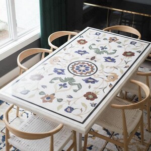 May include: A rectangular dining table with a white marble top, adorned with floral patterns in blue, red, and green. The table is surrounded by six wooden chairs with woven seats. A gold-toned circular light fixture hangs above.