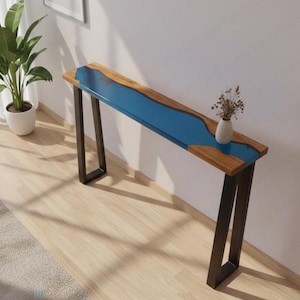 Puede incluir: Una consola de madera con un diseño de río de resina epoxi azul. La mesa tiene una parte superior rectangular con un centro azul y bordes de madera natural. Se apoya en patas de metal negro. Un pequeño jarrón blanco con flores secas está sobre la mesa.